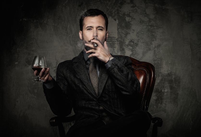 Stylishly dressed man in a dark suit, smoking a cigar and holding a glass of brandy while sitting in a leather char.